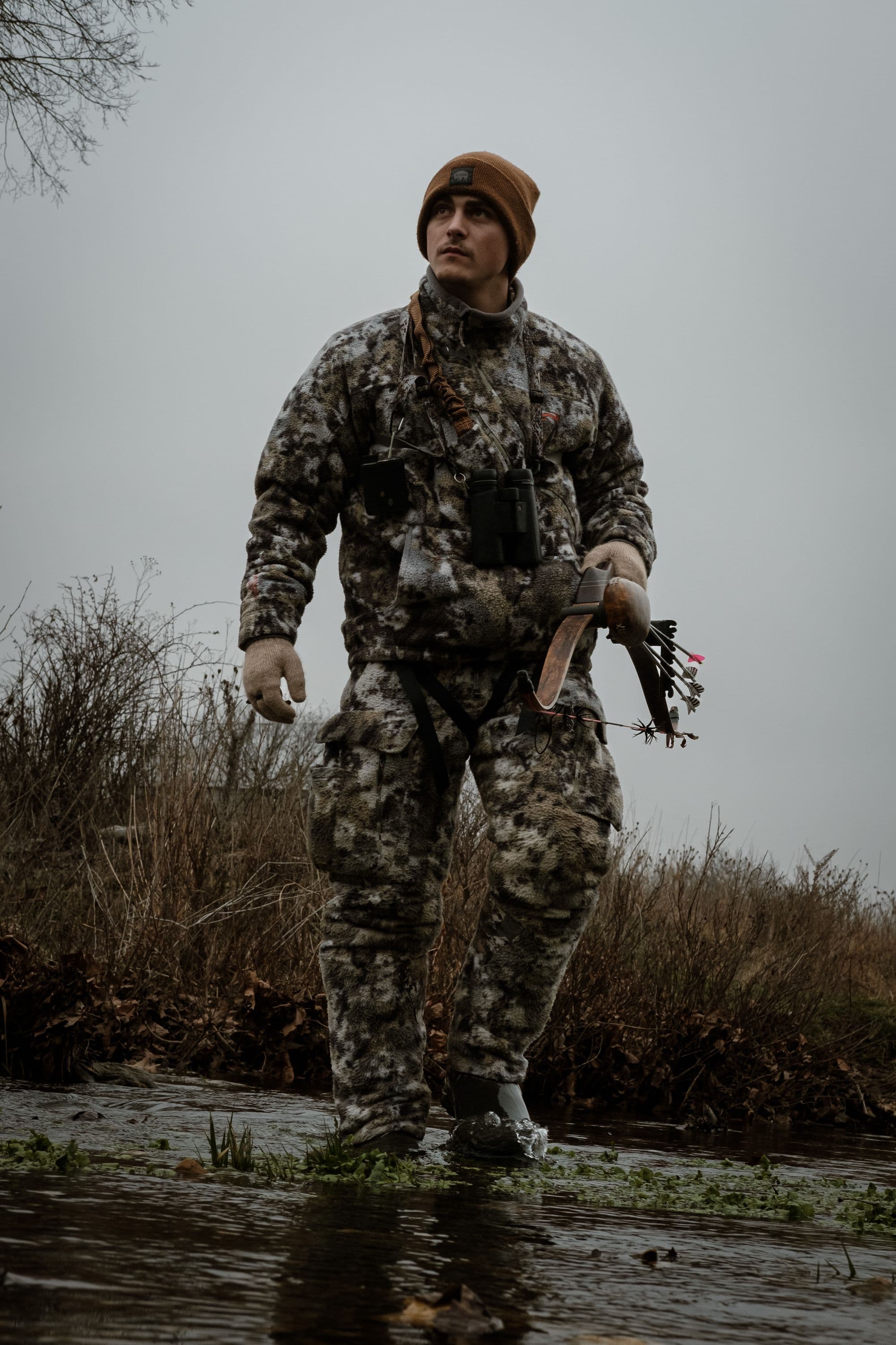 Bowhunter in camouflage crossing a river with a hunting bow