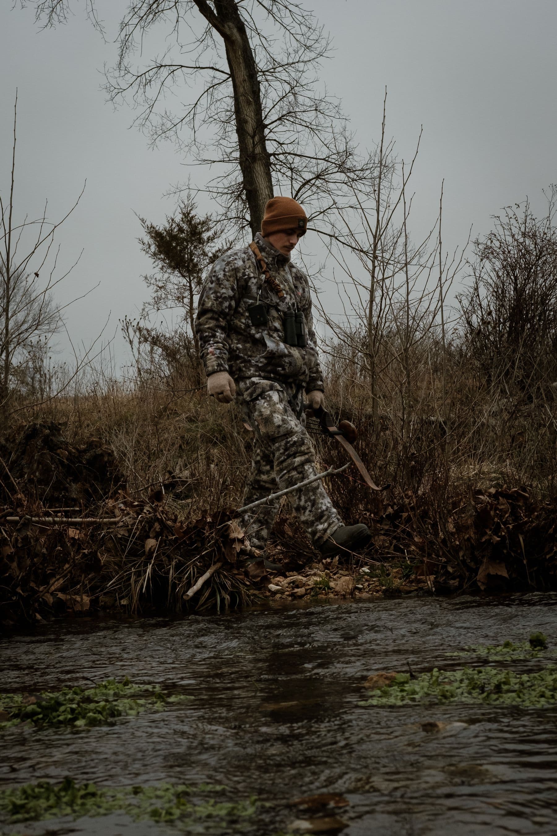 Bowhunter in camouflage walking a riverbank with a hunting bow